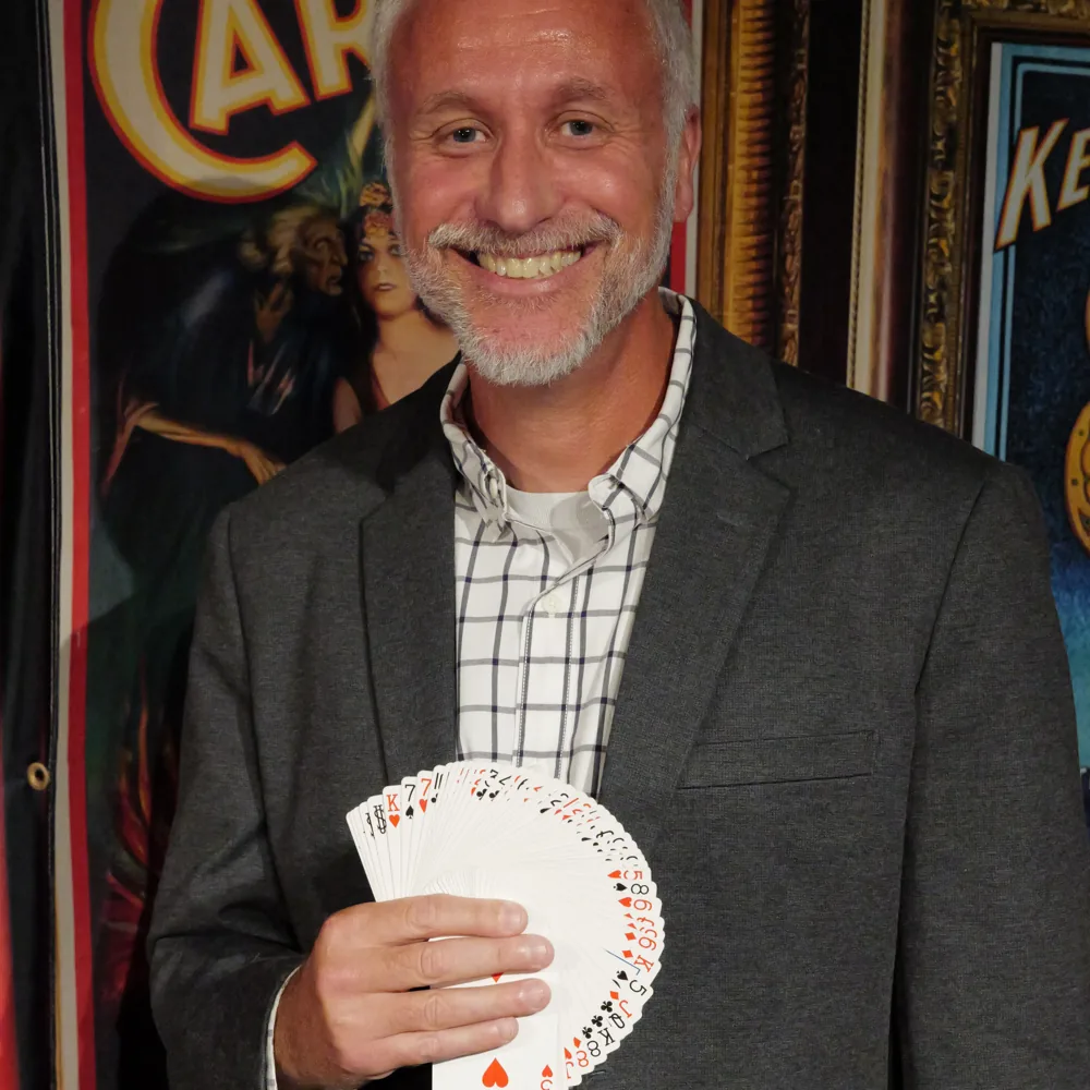 Mike Stringer smiling with a fan of playing cards in front of a vintage Carter magic poster
