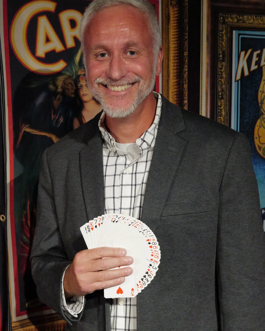 Mike Stringer smiling with a fan of playing cards in front of a vintage magic poster